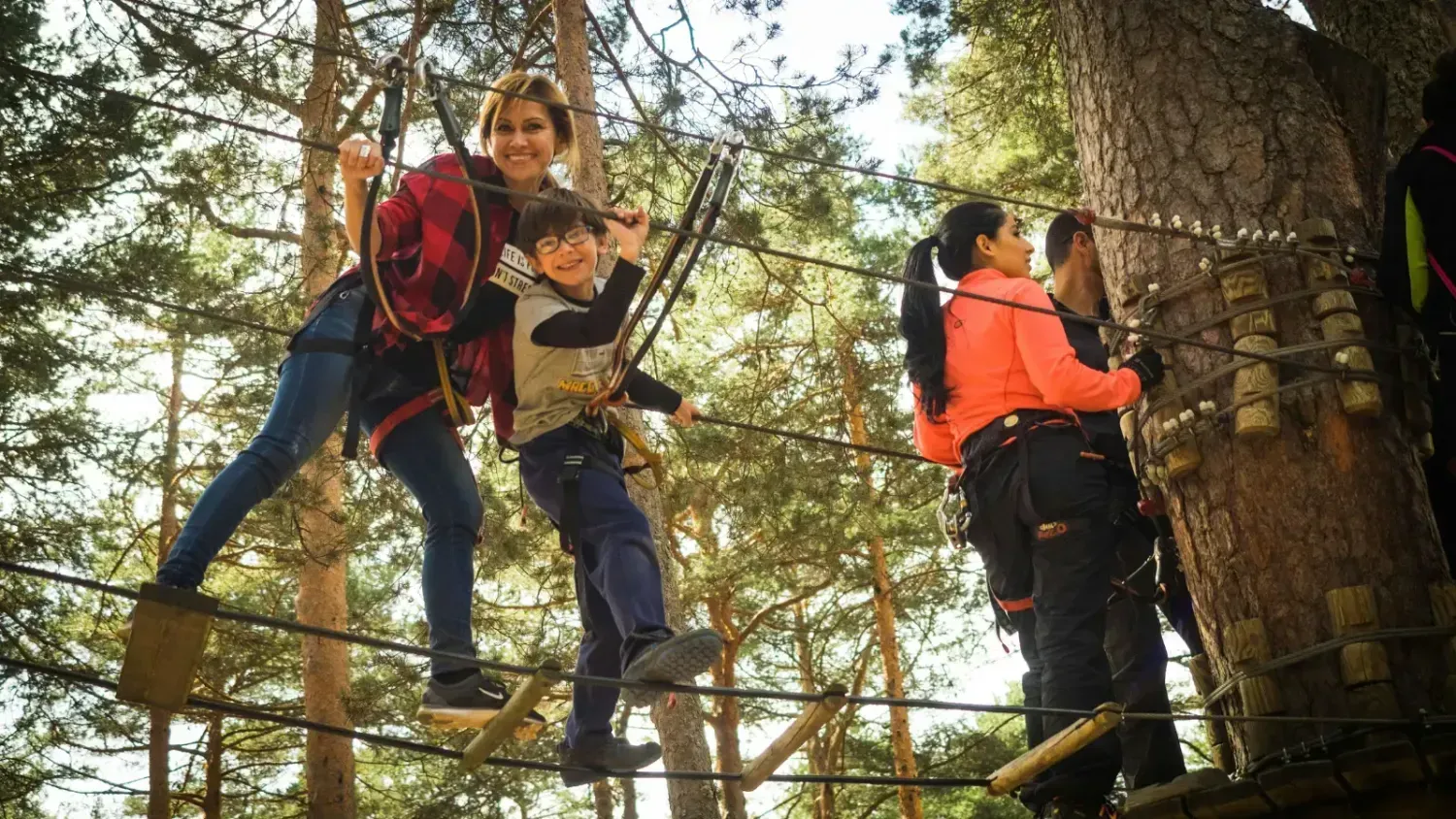 Family enjoying an outdoor adventure park with safety harnesses, mom and child smiling on rope bridge, representing family-friendly activities.