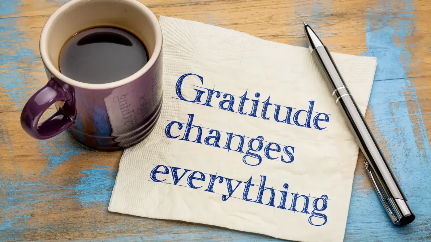 Purple coffee cup and silver pen beside a napkin with the words “Gratitude changes everything” on a rustic wooden table.