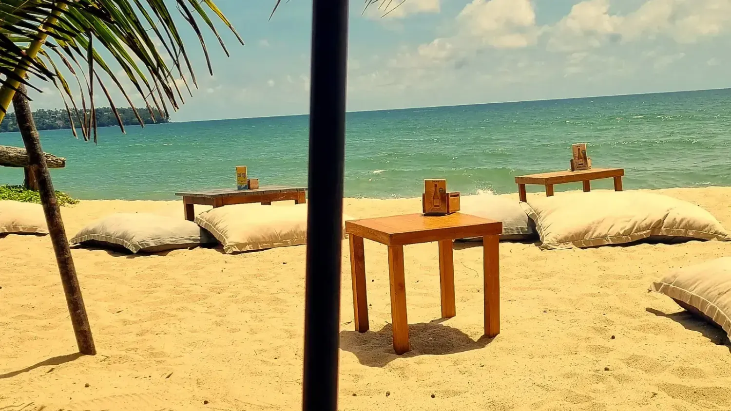 Wooden tables and beige floor cushions on a sandy beach facing the Andaman Sea in Bang Tao, Smigos Phuket, Thailand.