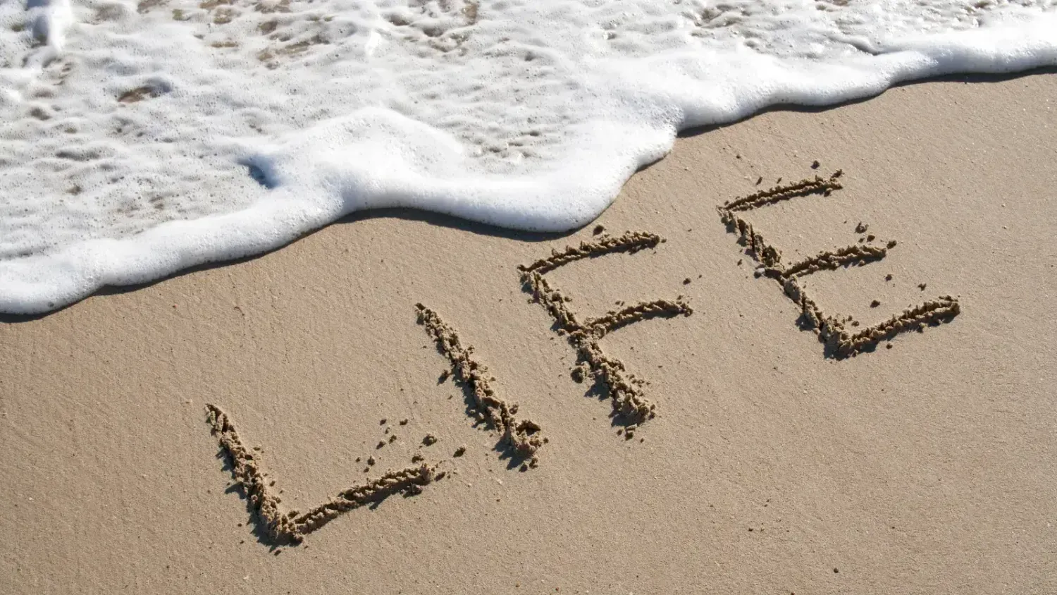 Word “LIFE” written in the sand with gentle sea foam approaching on a sunny beach in Phuket, Thailand.