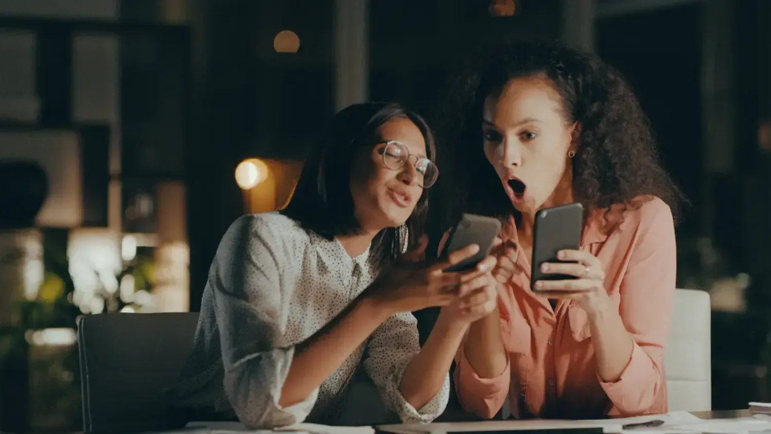 Two women looking at their phones together, reacting with excitement and surprise.