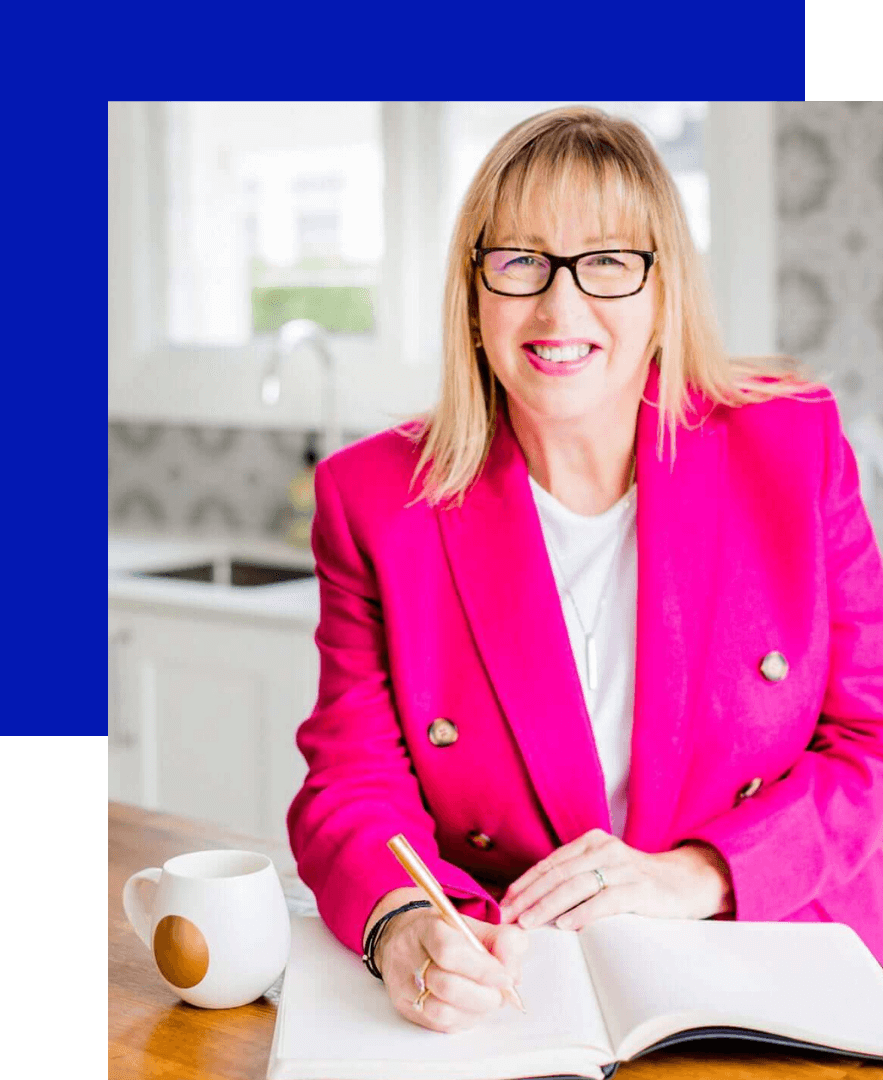Justine McLean, a woman with long blond hair, wearing glasses, a white shirt, and bright pink jacket, writing in a book at the kitchen counter, smiling at the camera	