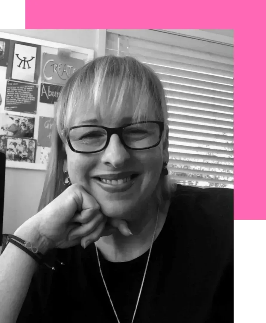 Black and white image of Justine McLean, a woman with long blond hair, wearing a dark shirt and glasses, in the office, smiling at the camera, resting her chin on her hand	