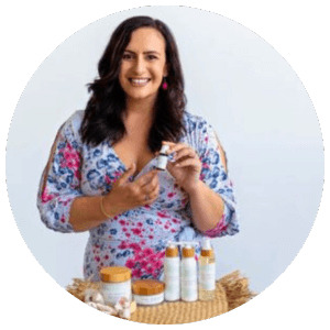 Natalie Kessell, a woman with long dark hair, wearing a floral dress, holding a glass bottle, in a kitchen, smiling at the camera	
