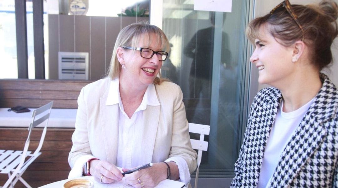 Justine McLean, a woman with long blond hair, wearing glasses and a white and cream shirt and jacket, sitting at a table with a client, a woman with short dark hair, wearing a checked jacket, smiling at the camer
