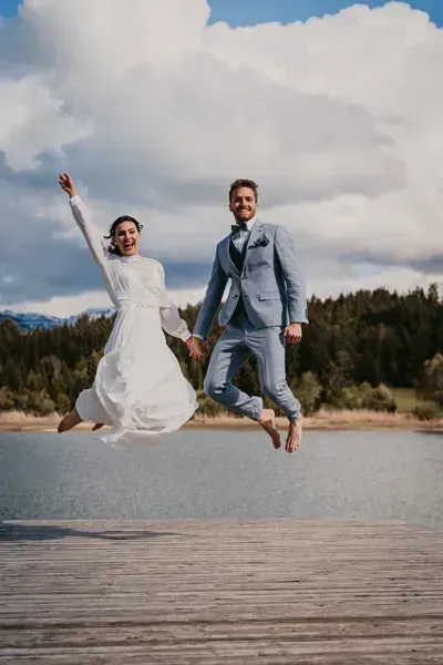 „Fröhliches Brautpaar in Hochzeitskleidung springt am Seeufer – emotionaler Hochzeitsmoment“