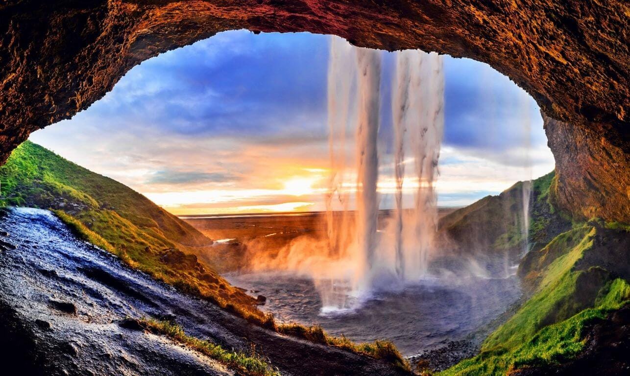Seljalandsfoss waterfall in Iceland seen from inside the cave-like overhang at sunset, with cascading water and lush green cliffs.