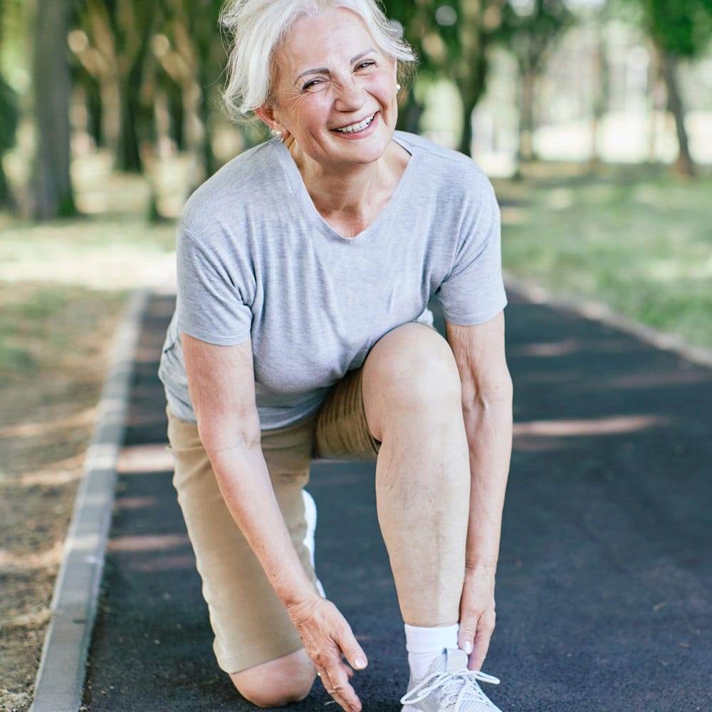 Smiling older woman crouching on a park path, one knee bent, hand at her ankle