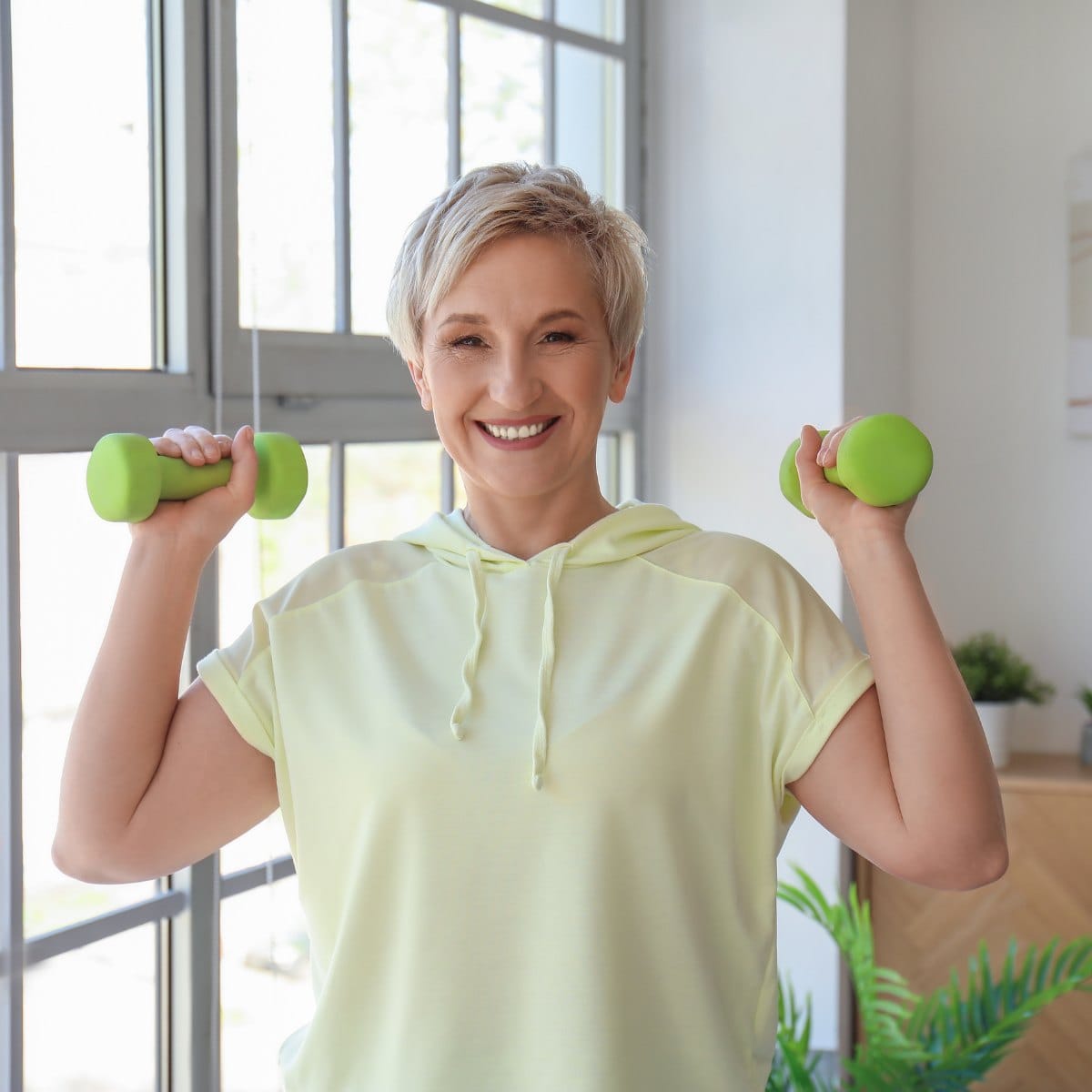 Smiling woman in her sixties holding a pair of dumbbells at home in a bright room