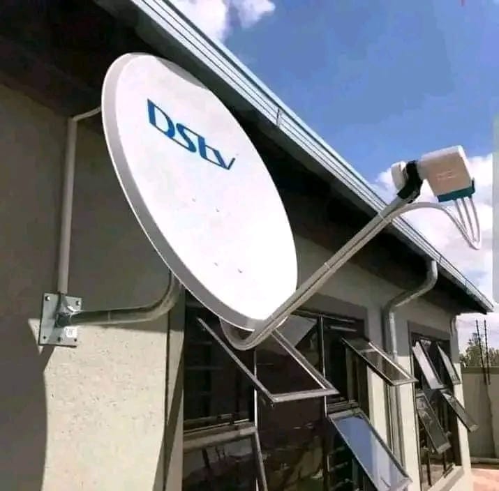 A white DStv satellite dish is mounted on the side of a building, positioned above several windows with security bars.