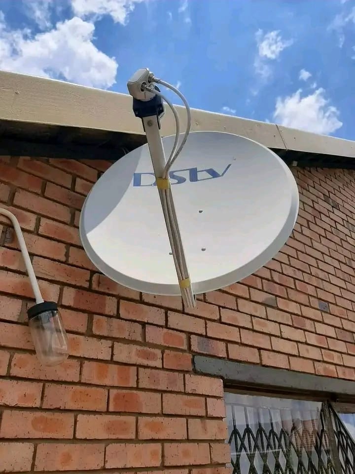 A white DStv satellite dish is mounted on a brick wall, with blue sky and clouds visible above.
