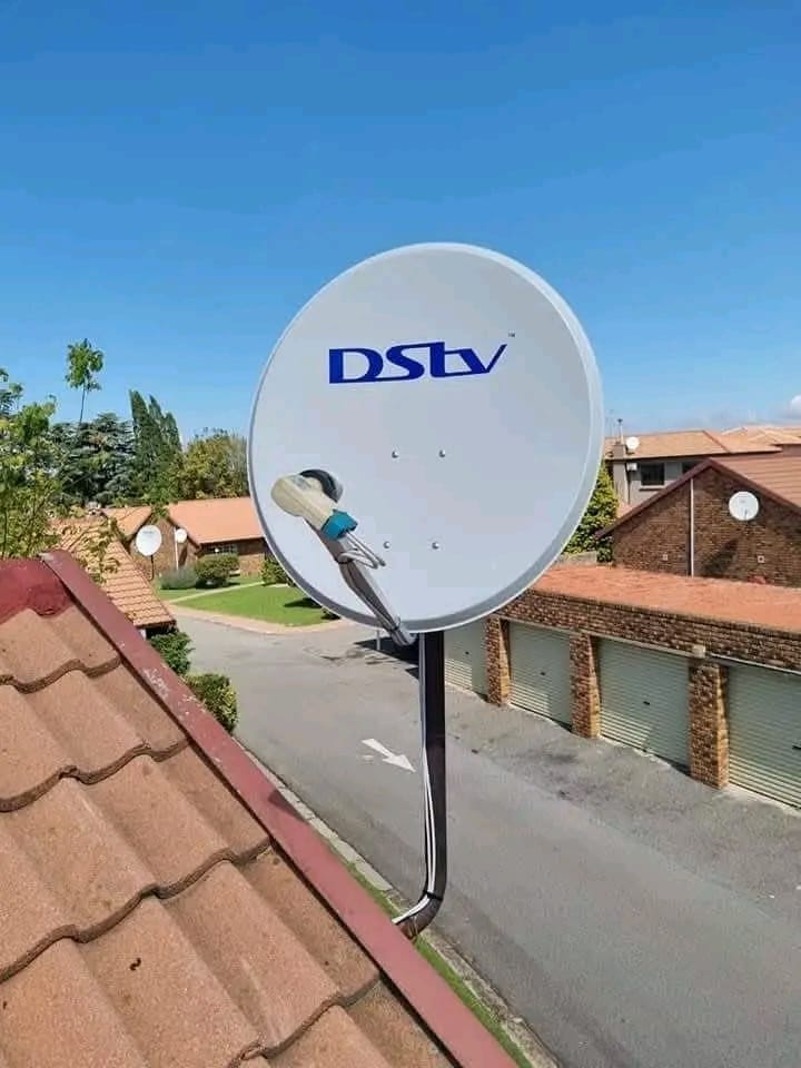 A DStv satellite dish is mounted on a pole, extending above a red-tiled roof with a residential street and houses in the background.
