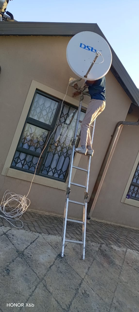 A person is standing on a silver ladder, appearing to work on a white DStv satellite dish mounted on the side of a light-colored building with windows protected by security bars. A coil of white cable is on the ground.