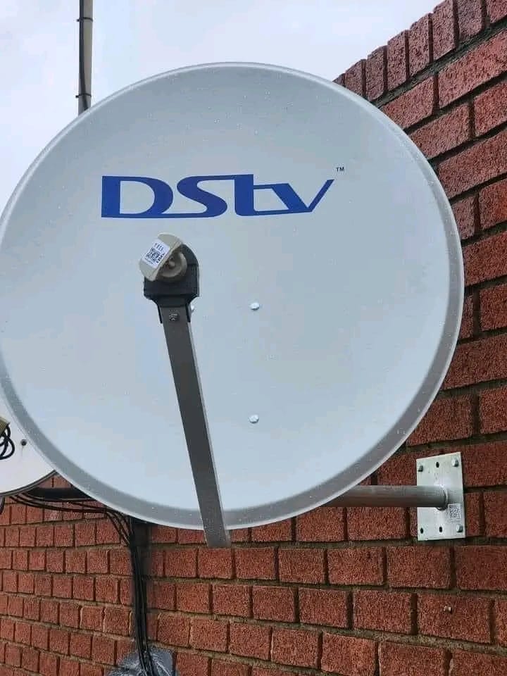 A white DStv satellite dish with blue "DStv" lettering is mounted on a reddish-brown brick wall.