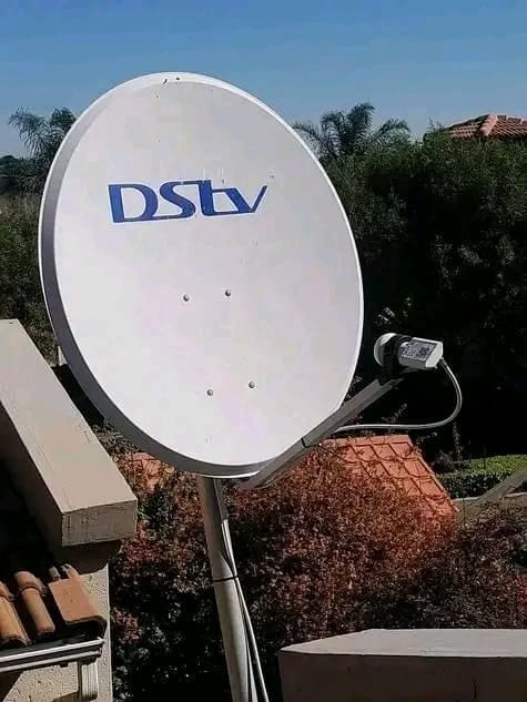 A white DStv satellite dish is positioned outdoors, with green trees and a tiled roof in the background under a blue sky.