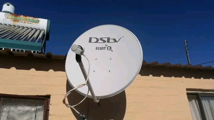 A DStv satellite dish is mounted on a yellow wall, with a solar water heater visible on the roof in the background under a clear blue sky.
