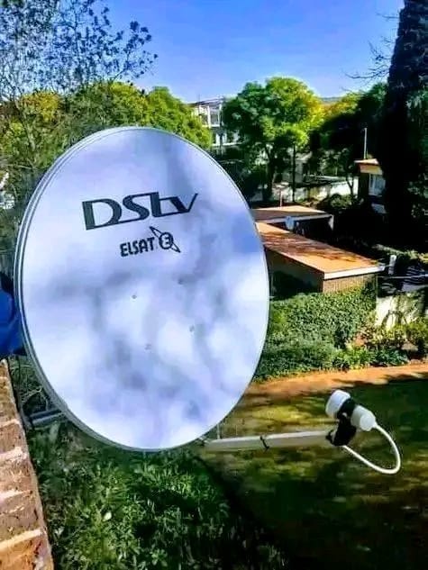 A DStv ELSAT satellite dish is visible outdoors with lush green trees and residential buildings in the background.
