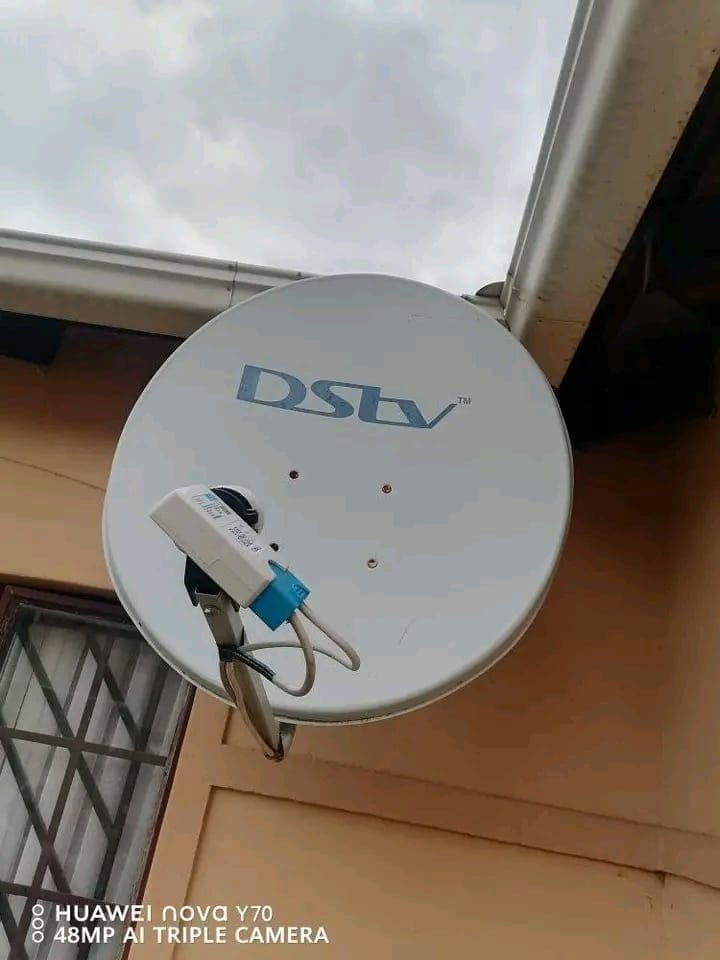 A white DStv satellite dish is mounted on a light grey exterior wall of a building, positioned below the gutter. Several open awning-style windows with security bars are visible beneath the dish.