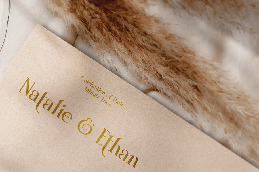 Close-up of a rustic wedding invitation card for Natalie and Ethan featuring elegant gold typography printed on textured kraft cardstock next to soft, fluffy pampas grass.