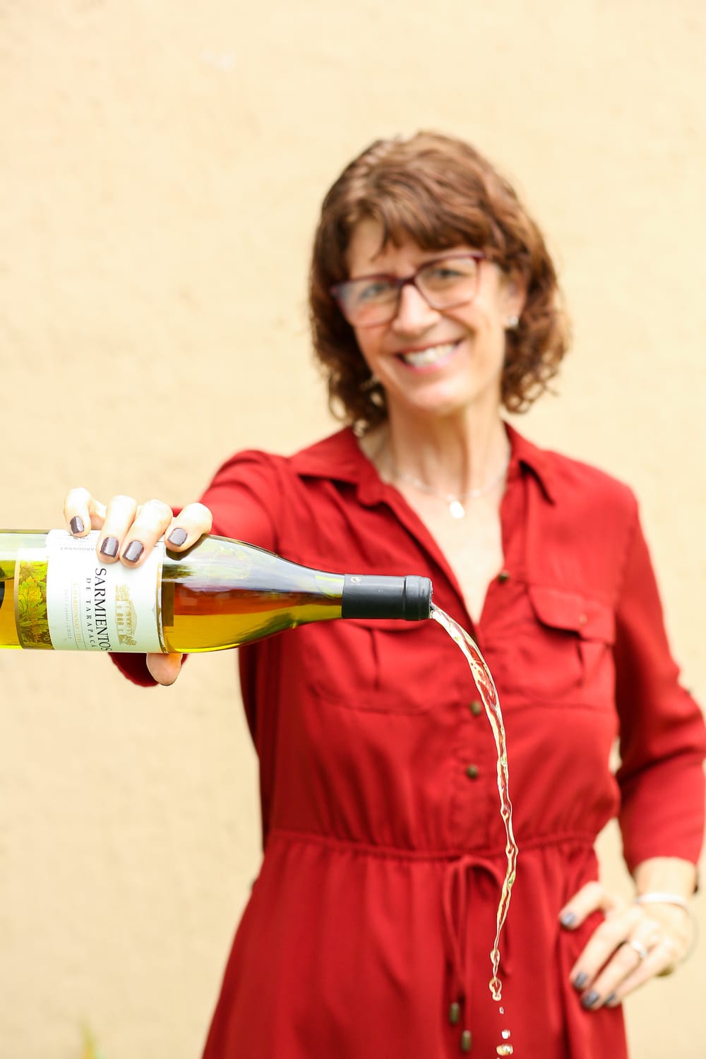 author pouring out a bottle of white wine. She is blurred in the background wearing a red dress, arm extended with wine bottle held horizontally
