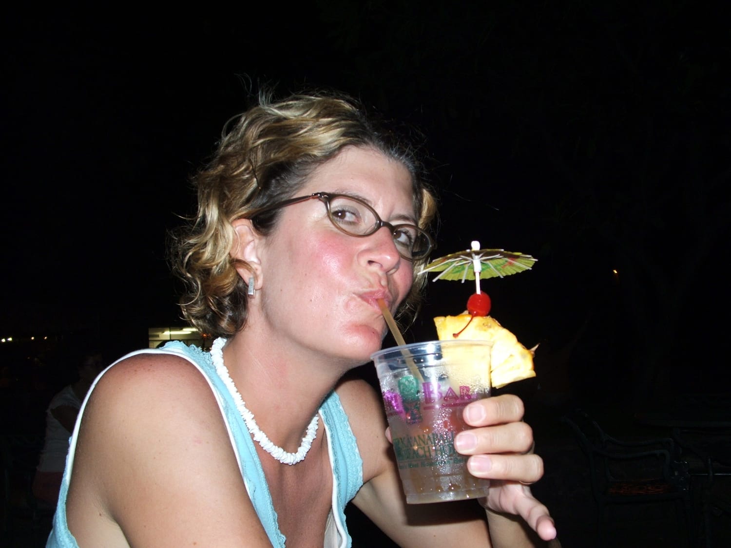 Author in 2005 wearing a light blue tank top and a puka shell necklace. Sipping a Mai Tai. Drink has a wedge of pineapple and a paper umbrella 