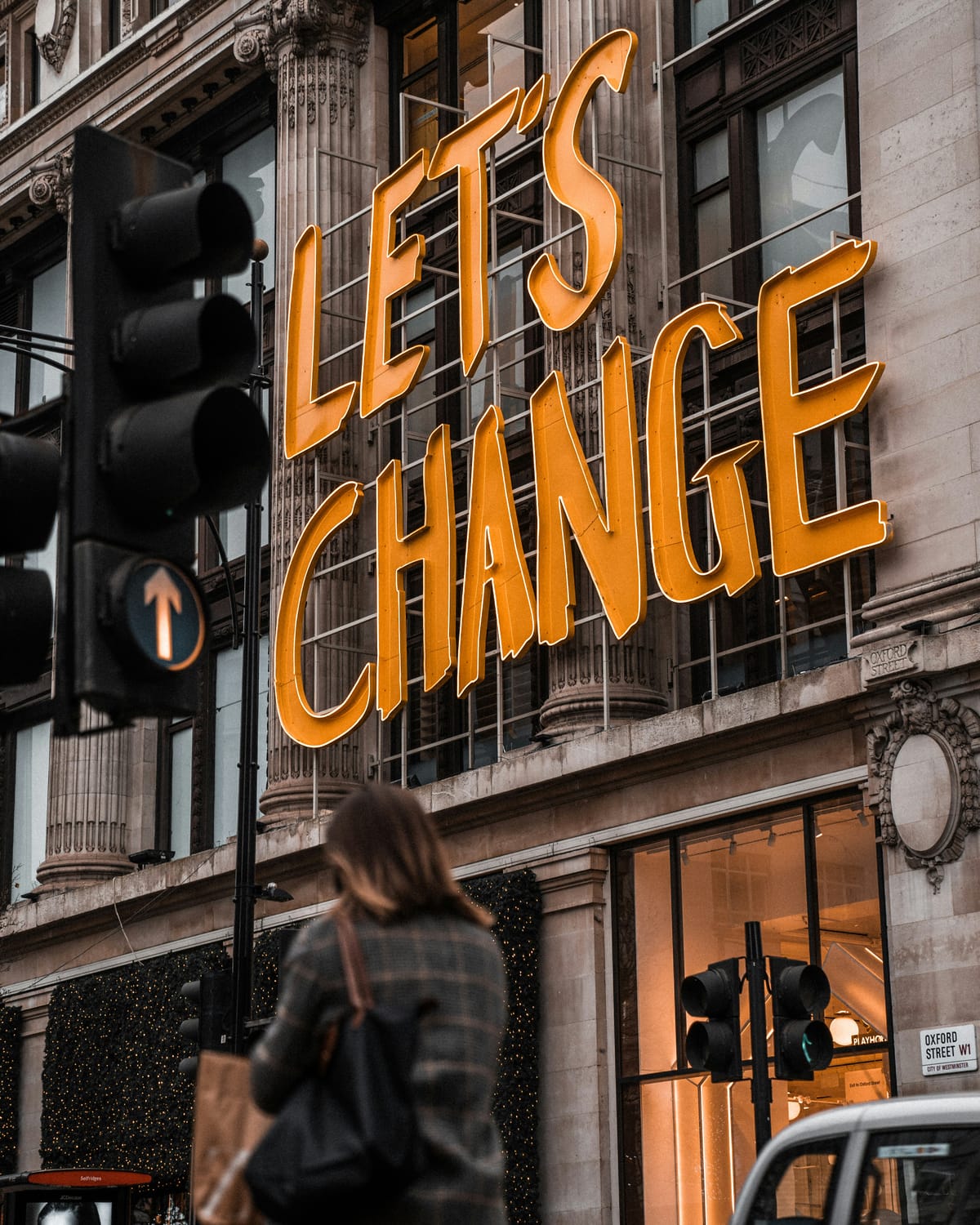 building in a downtown that has a large sign that saying "Let's Change". There is a blurred woman walking in front of it.