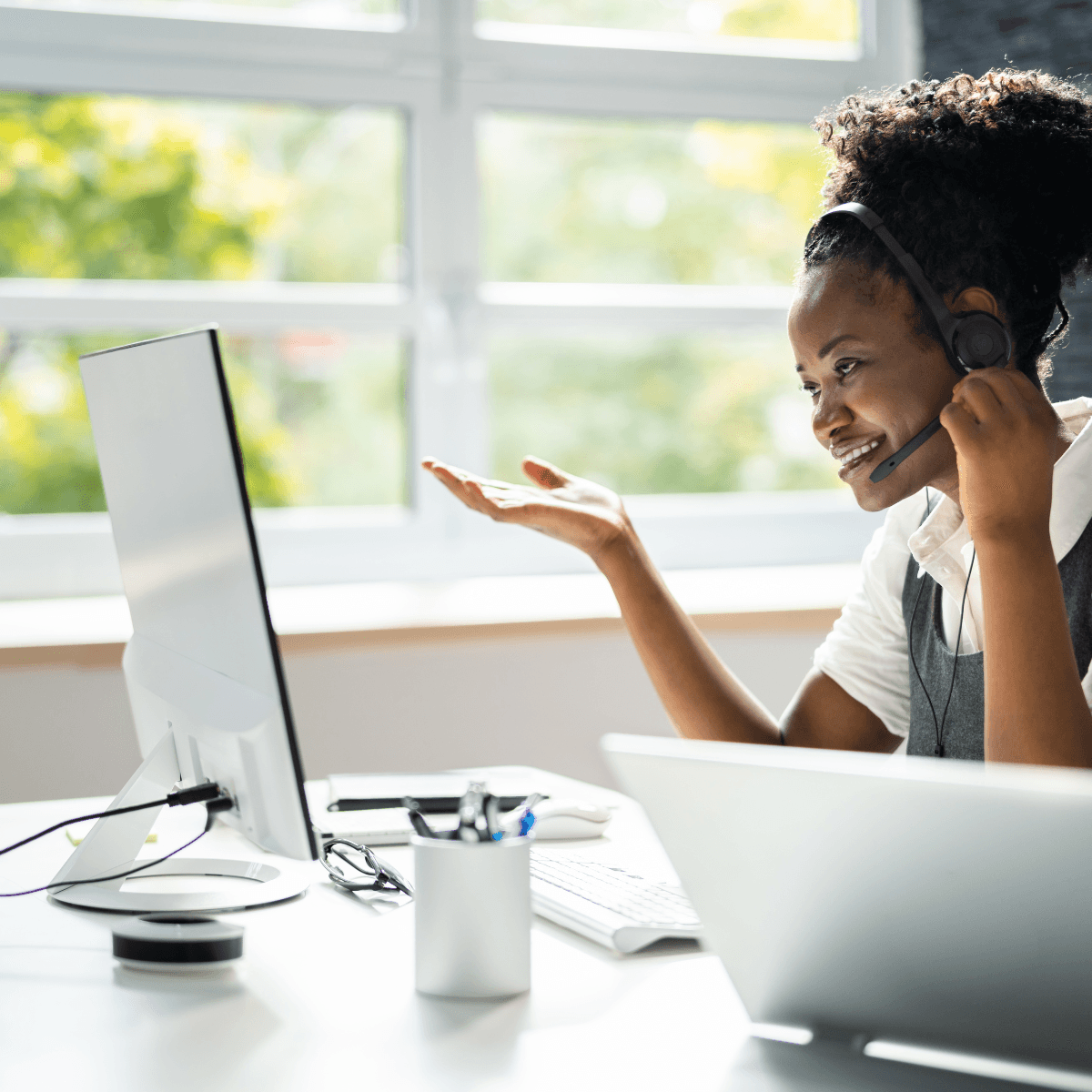 A smiling virtual assistant wearing a headset, gesturing to a computer screen while working, representing professional virtual assistance services including social media marketing, customer support, and email management.