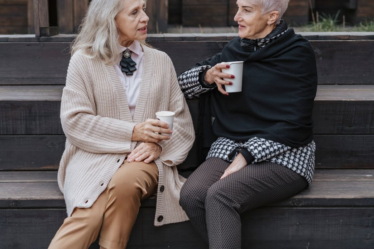 Dwie starsze kobiety siedzą obok siebie z kubkami i rozmawiają