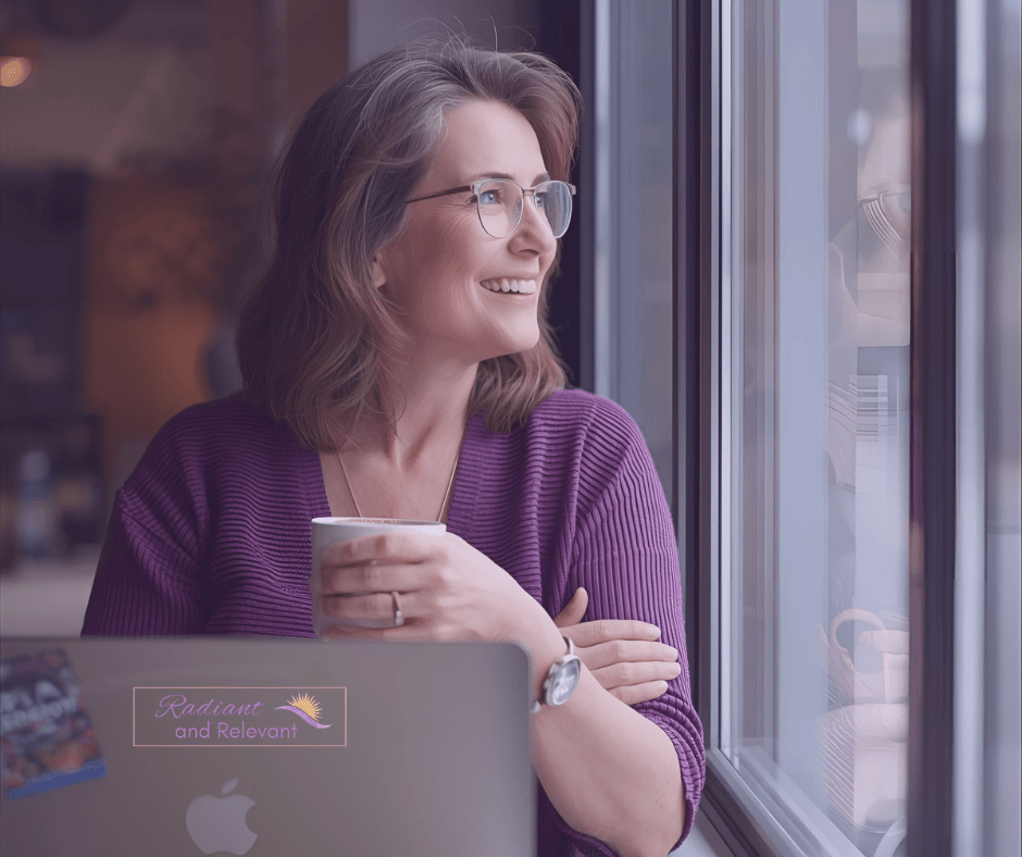 women holding coffee cup looking out window smiling with laptop on table in front of her