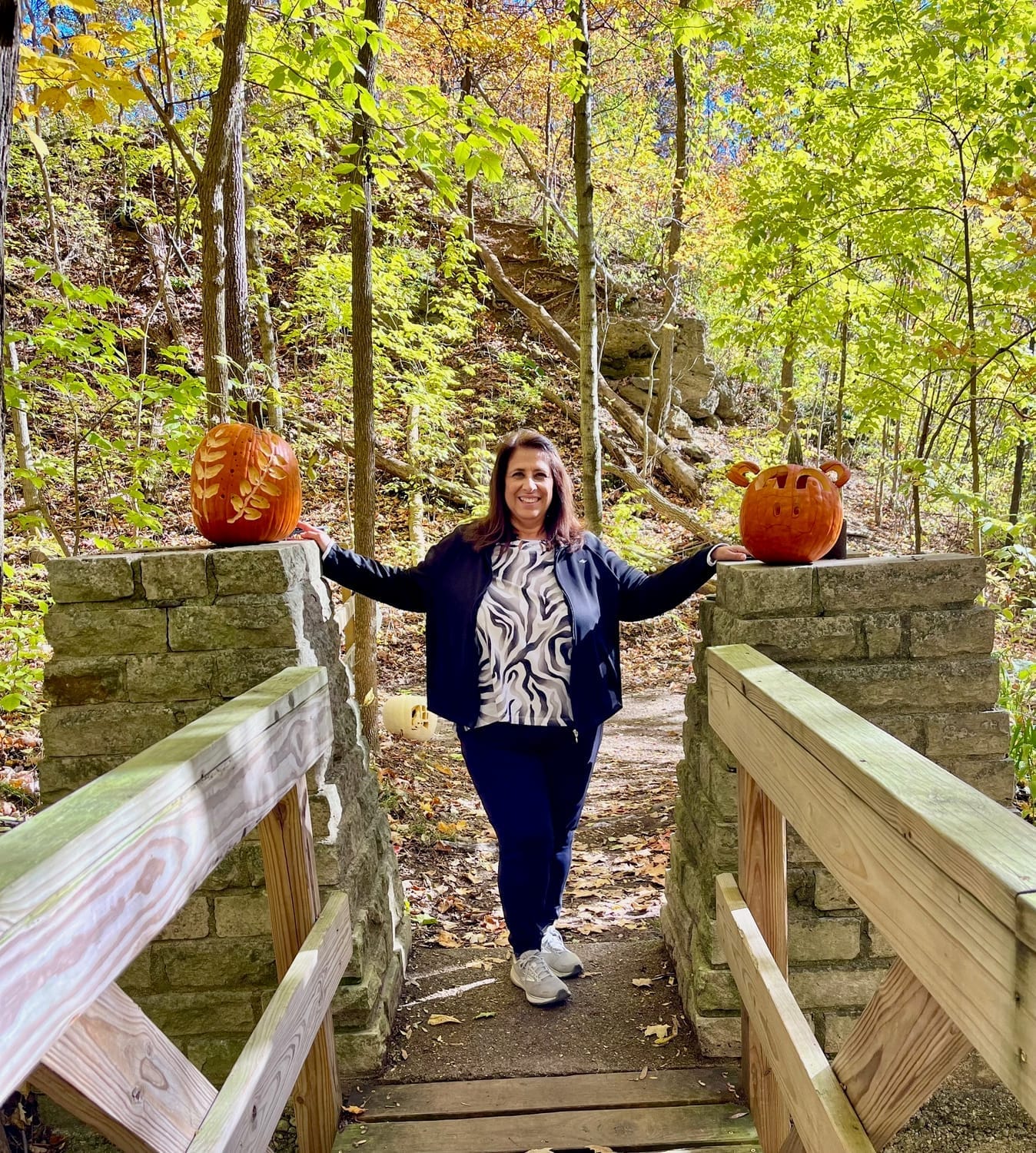 janet farmer standing outdoors near fall foliage and at the end of a bridge with 2 pumpkins
