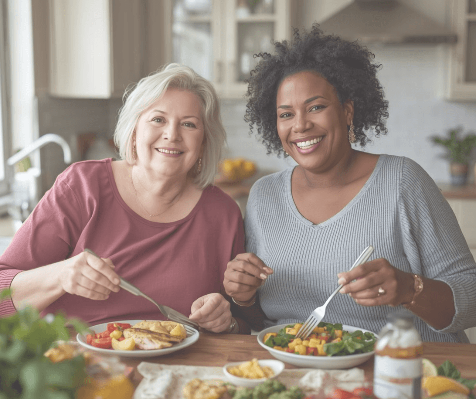 a white and a black woman aged 50-60 smiling and eating a health meal together in their kitchen