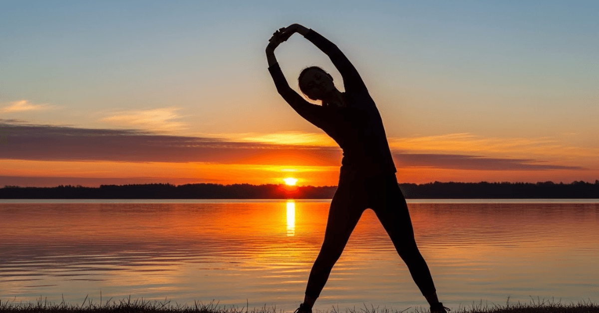 Silhouette of a person stretching by the water at sunset, symbolizing how consistent daily movement supports long-term health, energy, and longevity.