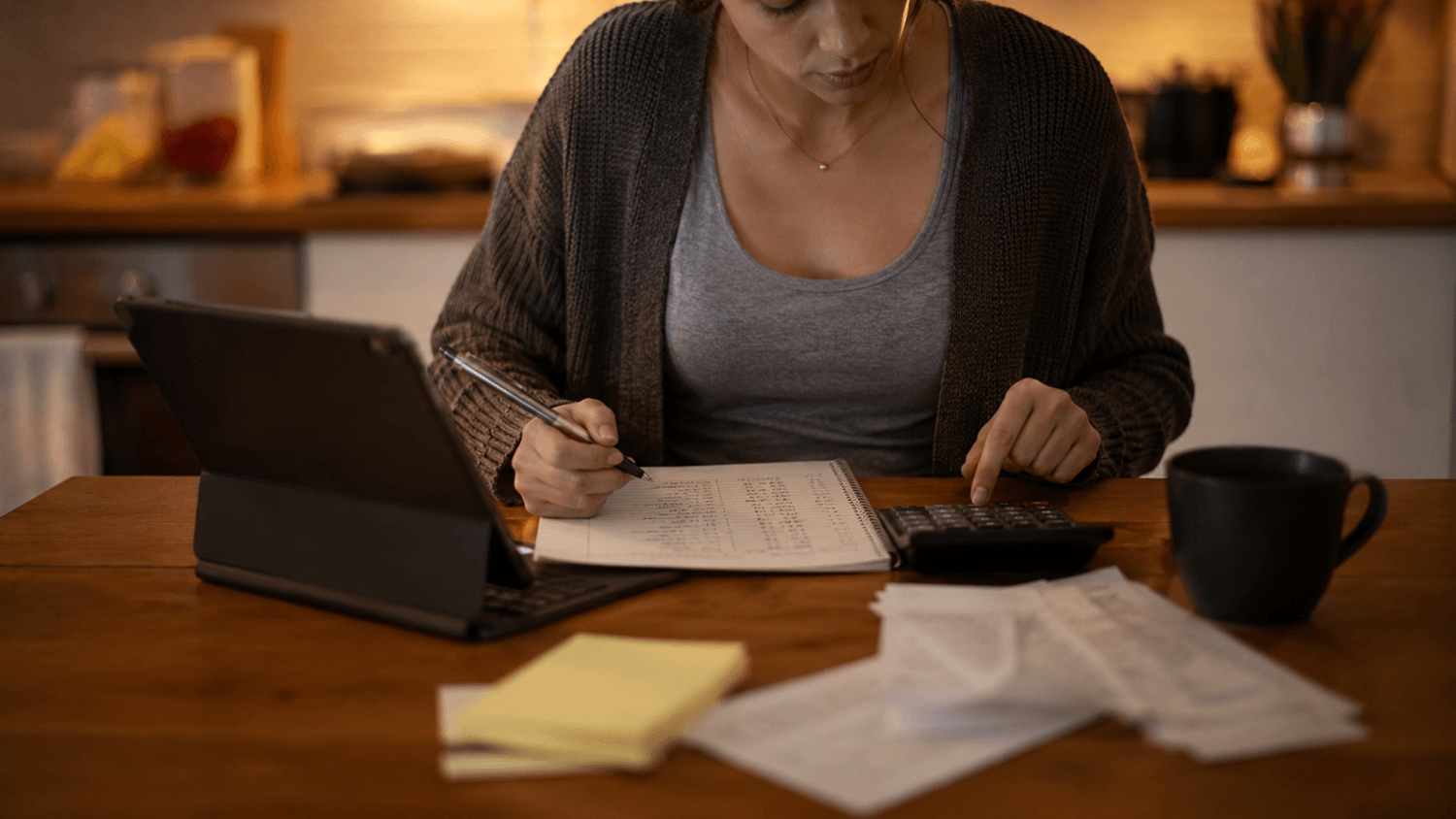 Person using a tablet, calculator, and budget sheet to organize monthly expenses at a kitchen table.