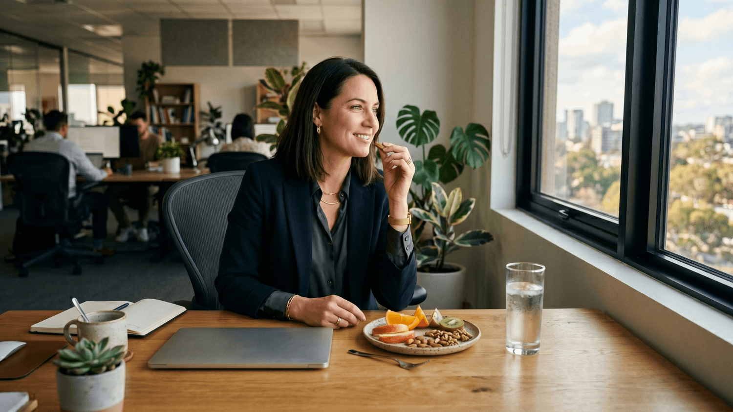 Professional enjoying a balanced afternoon snack with fruit, nuts, and water beside a large office window.