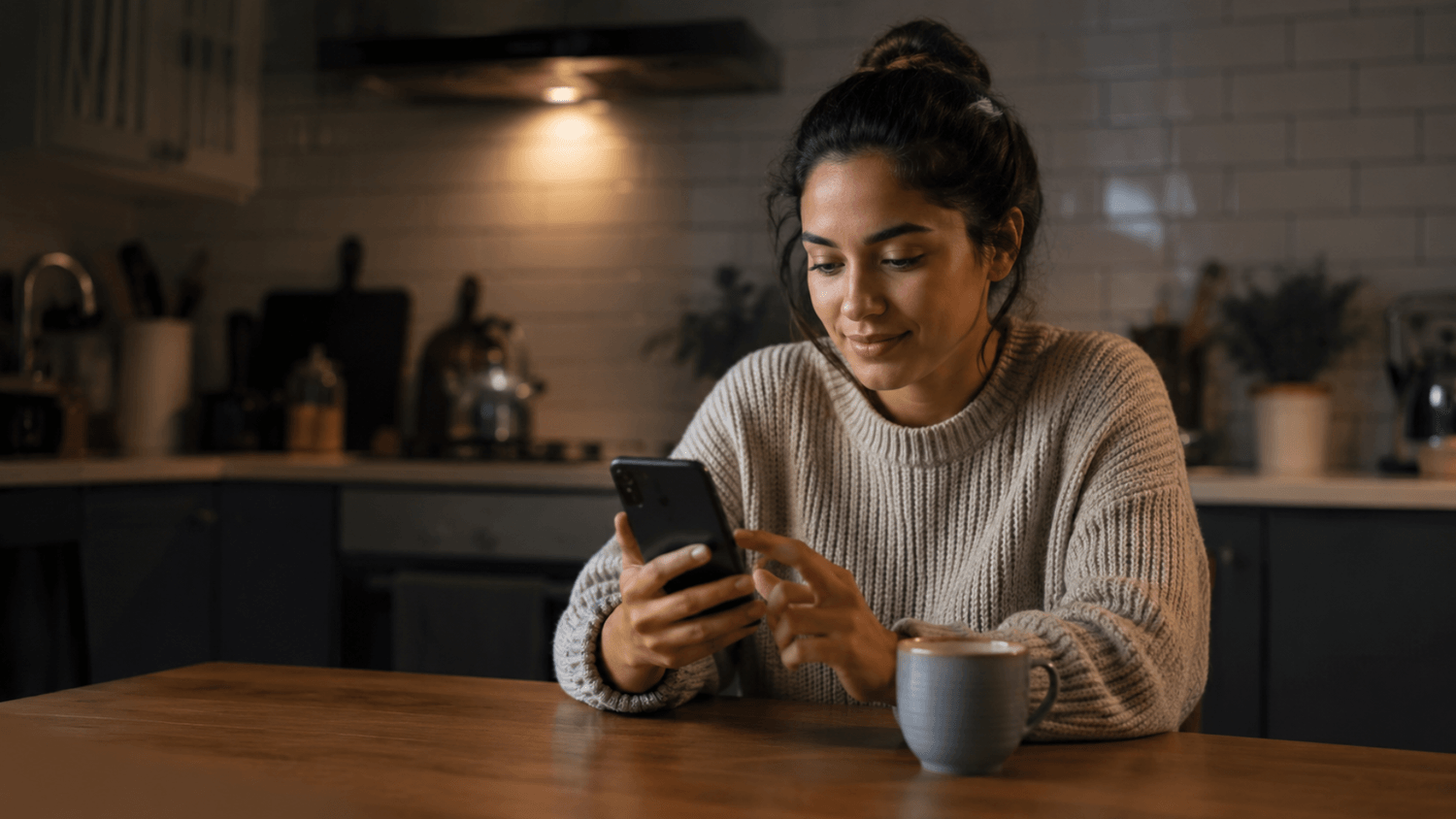 Person using a smartphone at a kitchen table to post or share content online in a relaxed evening setting