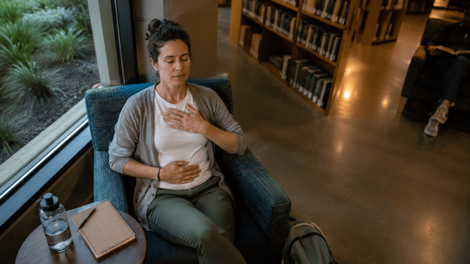 Adult completing a quiet body check-in beside a notebook and water bottle in a library seating area.
