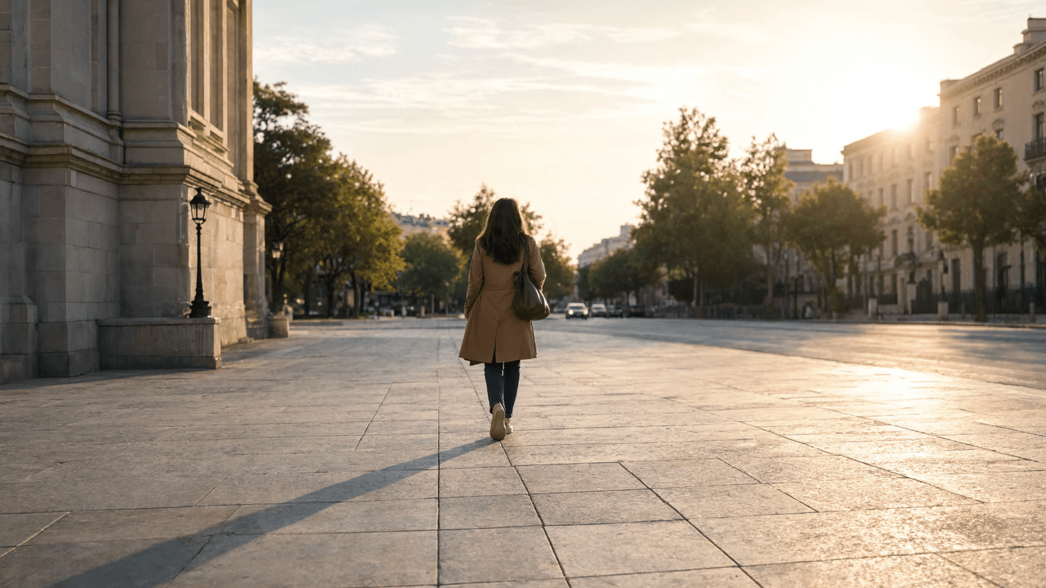 Adult walking confidently across an open public plaza at sunrise toward a tree-lined street.