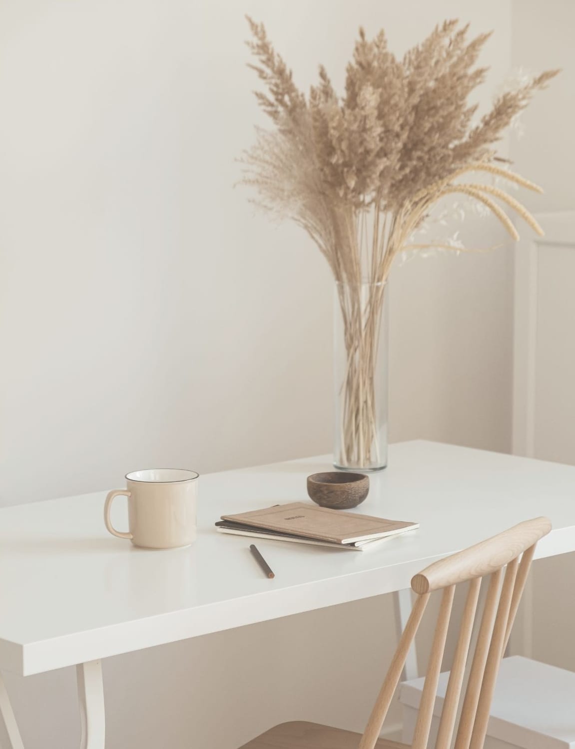Minimalist white desk with notebook, mug, and vase of dried grasses — calm workspace aesthetic for focus and reflection.
