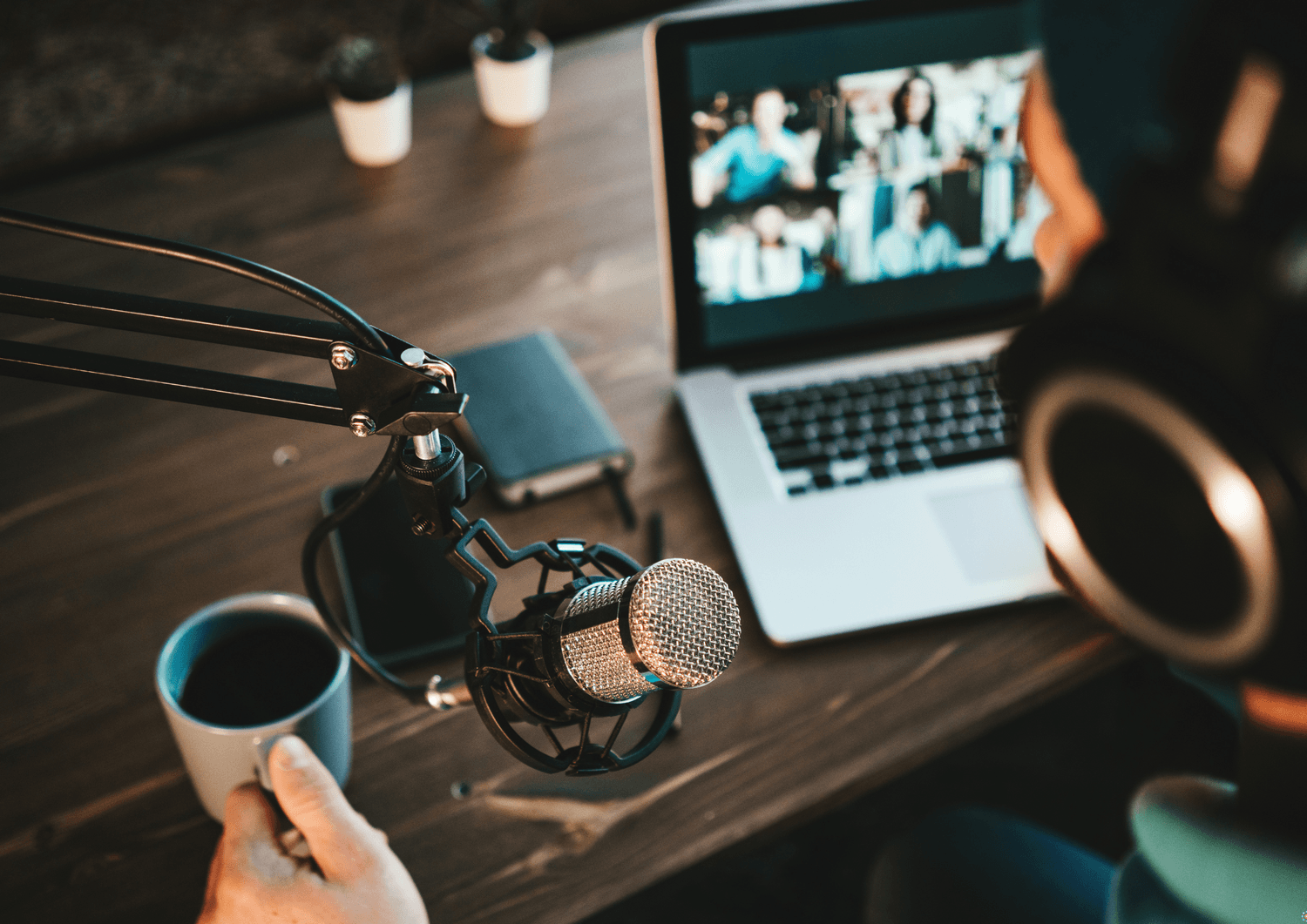 Person med kondensatormikrofon og hodetelefoner ved et skrivebord, med en laptop som viser et videomøte i bakgrunnen – illustrerer digital innholdsproduksjon