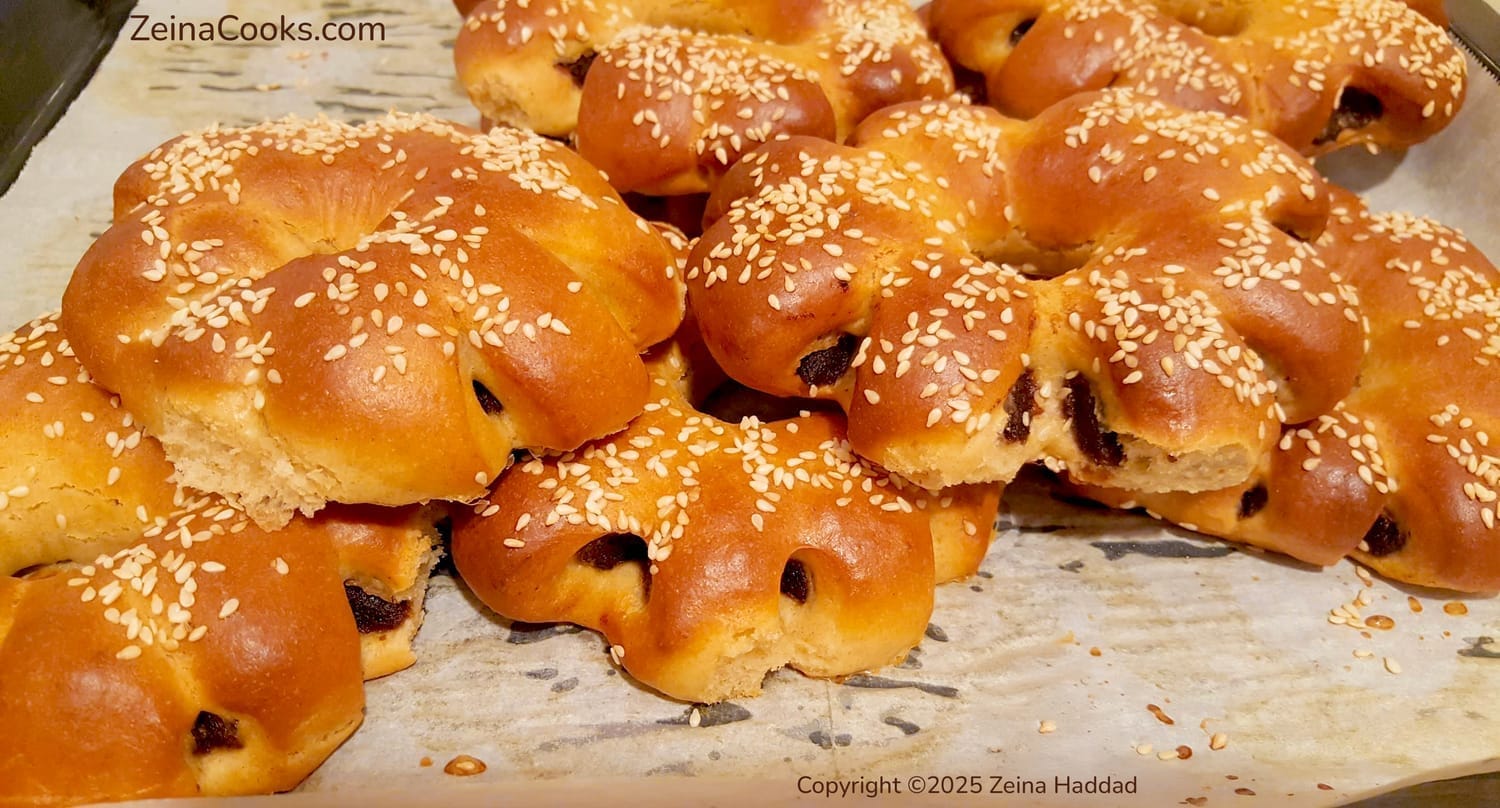 Close-up of Golden brown Arabic brioche rings filled with dates and topped with sesame seeds on a baking sheet freshly out of the oven by ZeinaCooks.com