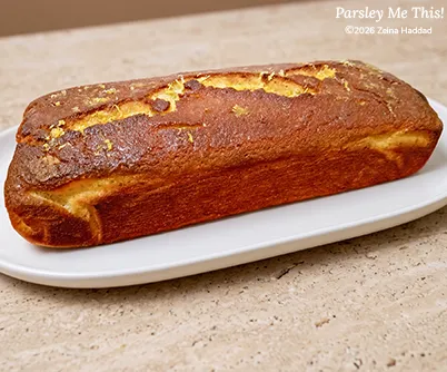 The cake "Zesty Lemon Butter Cake With Glaze" by Parsley Me This, having been baked in a loaf pan, resting on a white platter.