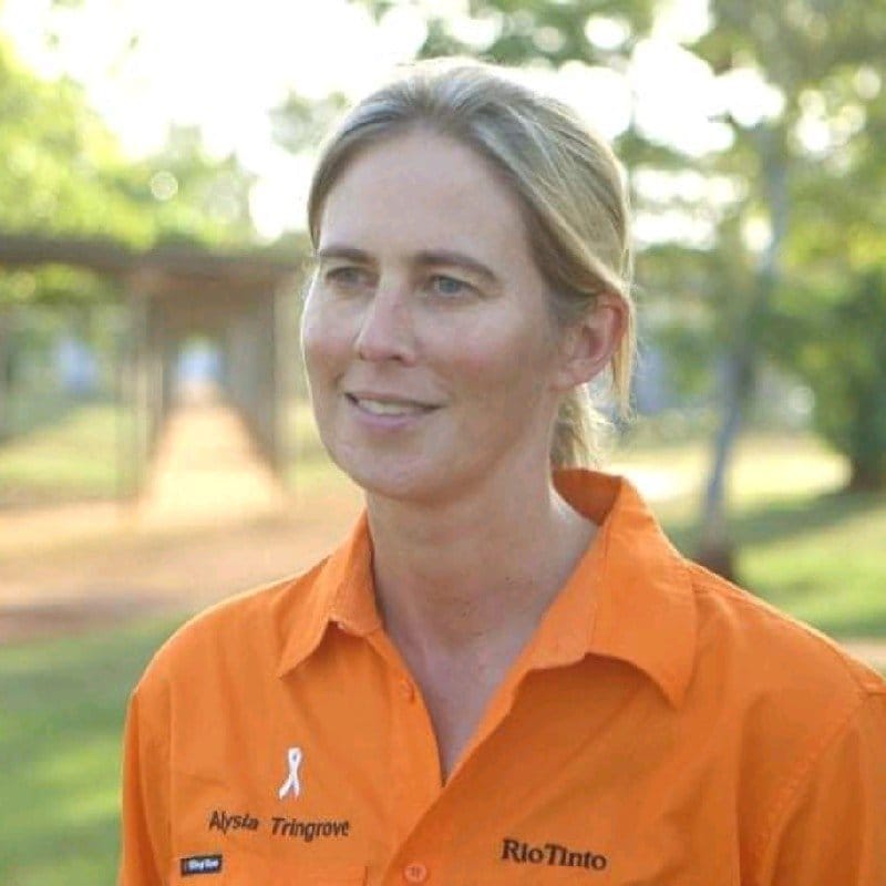 A woman in an orange polo shirt standing outdoors with trees and a platform in the background.