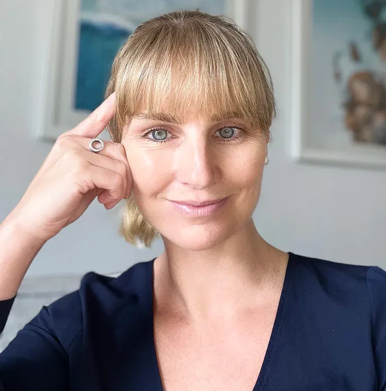picture of laura thain, blonde hair tied back in ponytail, fringe, navy v neck top smiling to camera