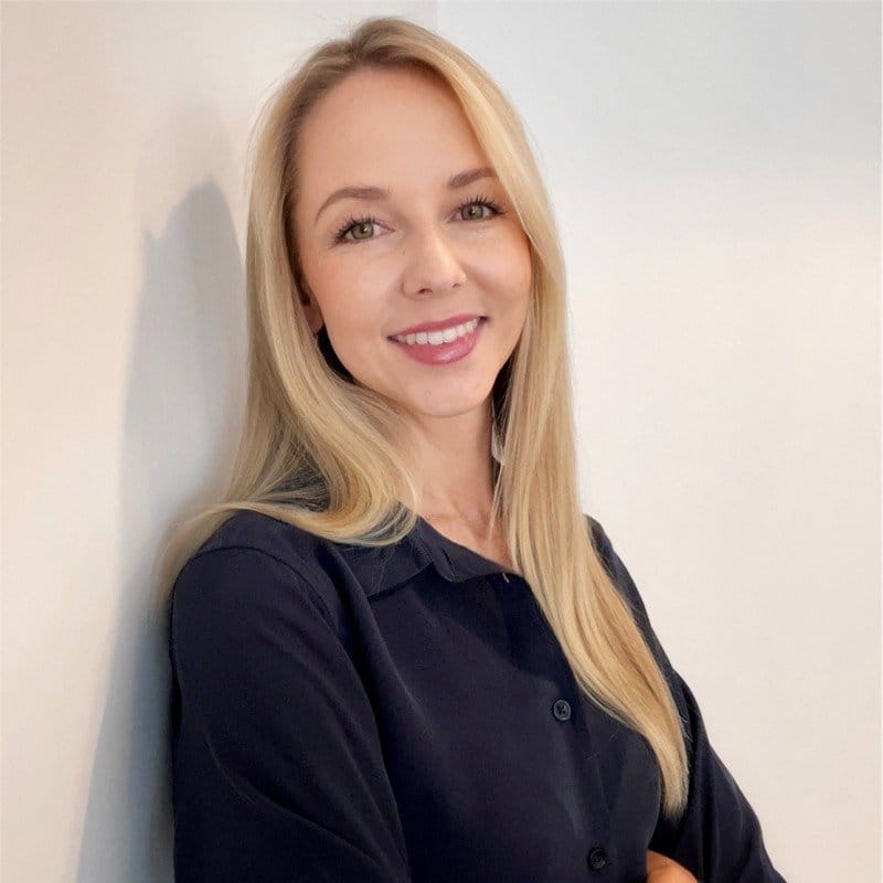 A blonde woman in a black shirt smiling and leaning against a white wall.