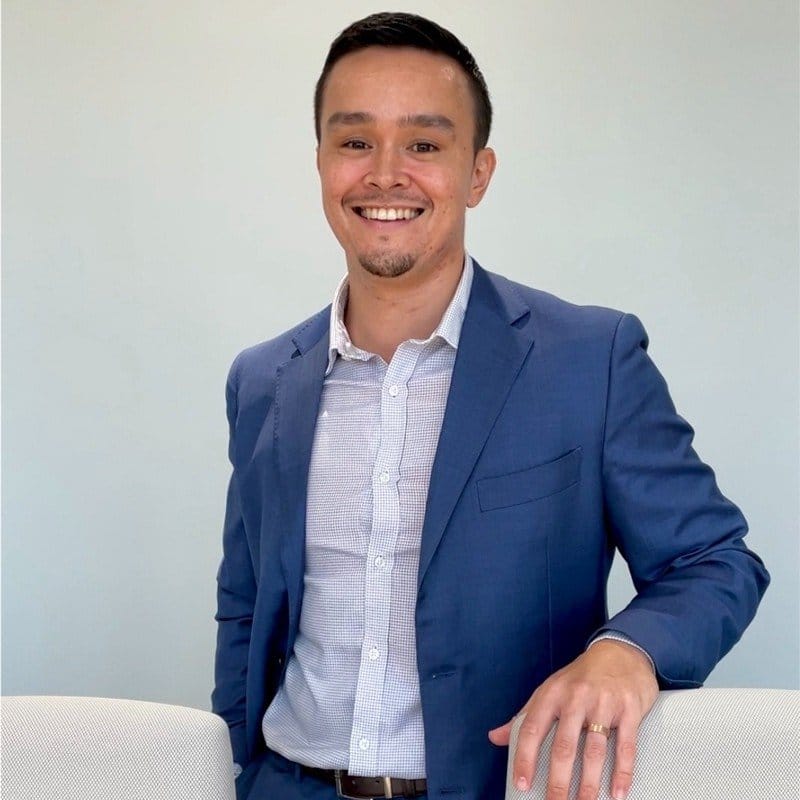 A smiling man in a blue suit with a light-colored dress shirt standing indoors against a plain wall.