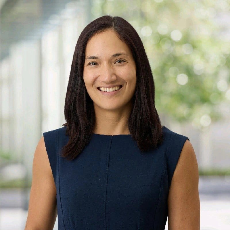 A woman with long dark hair, wearing a sleeveless navy blue top, standing outdoors with blurred greenery in the background, smiling at the camera.