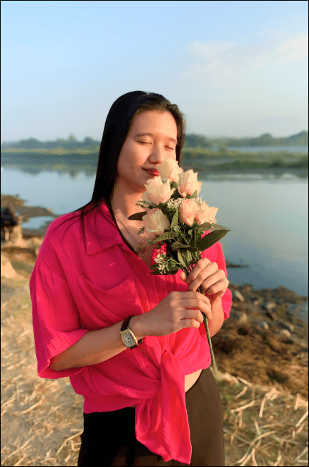 Love Luna in a bright pink blouse stands near a riverbank, holding a bouquet of pale pink roses close to her face with her eyes closed, enjoying the flowers.