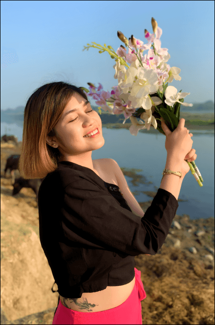 Ella Luna in a black outfit smiles with her eyes closed while holding a bouquet of white and purple orchids, standing by the riverside in the sunlight.