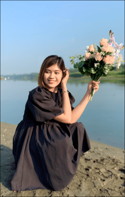Ella in a black outfit sits by the riverside, smiling while holding a bouquet of light pink roses.
