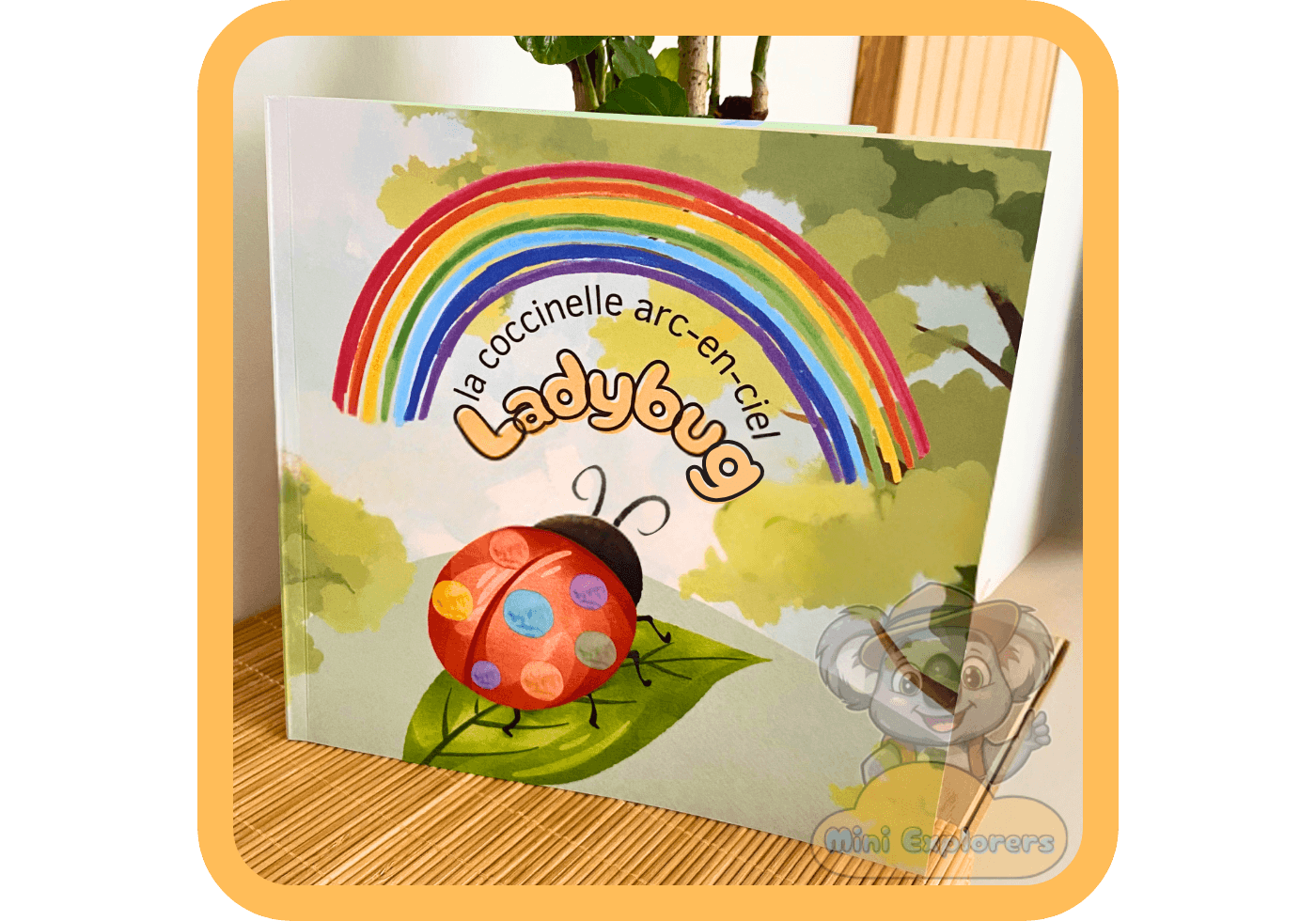 Une photo de la couverture du livre pour enfants "la coccinelle arc-en-ciel Ladybug", présentée dans un cadre numérique orange clair. La couverture illustrée montre une grande coccinelle rouge avec de multiples points de couleurs différentes (bleu, vert, jaune, etc.) sur son dos, assise sur une feuille verte. Au-dessus d'elle se dresse un grand arc-en-ciel coloré, et le fond est un paysage de jardin flou avec des arbres et de la verdure. En bas à droite, il y a un filigrane numérique du personnage de koala explorateur de Mini Explorers.